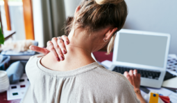 Woman seen from behind at a laptop, hand pressed to the back of her neck as she tilts her head forward.